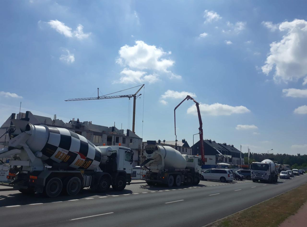 Kaskada betonomieszarek w ruchu drogowym na tle budowy z żurawiem i pompą do betonu, błękitne niebo z obłokami.