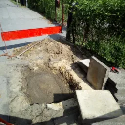 Wykop w chodniku zabezpieczony czerwoną barierką, obok stos betonowych płyt i narzędzie, widok na ulicę i zieleń.