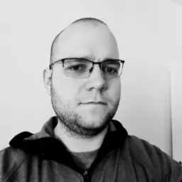 Czarno-białe zdjęcie portretowe mężczyzny w okularach i bluzie z kapturem, z krótką brodą, na tle jasnej ściany. Wyraz twarzy poważny, ujęcie z bliska.