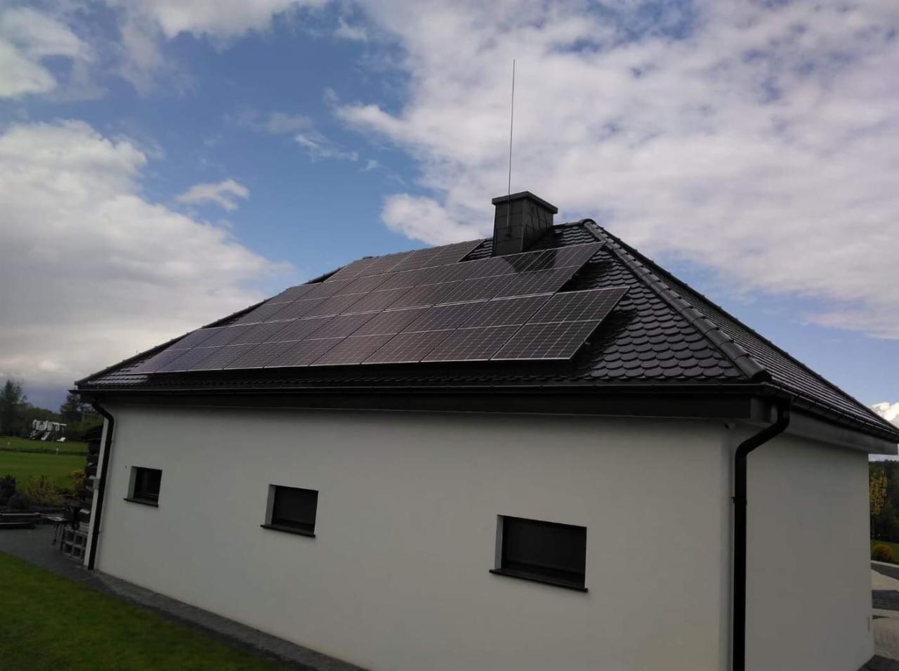 Panele fotowoltaiczne na dachu domu jednorodzinnego z czarną dachówką i piorunochronem, widok z dołu na tle błękitnego nieba z chmurami.