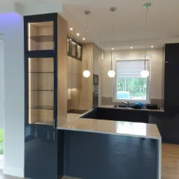 Nowoczesna kuchnia z wyspą, oświetleniem punktowym i przeszkloną witryną z podświetleniem LED, fronty drewnopodobne i czarne, jasny blat.