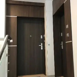 Dwa wejścia do mieszkań w bloku, ciemne drzwi z zabudową i srebrnymi akcentami, wycieraczki przed drzwiami.