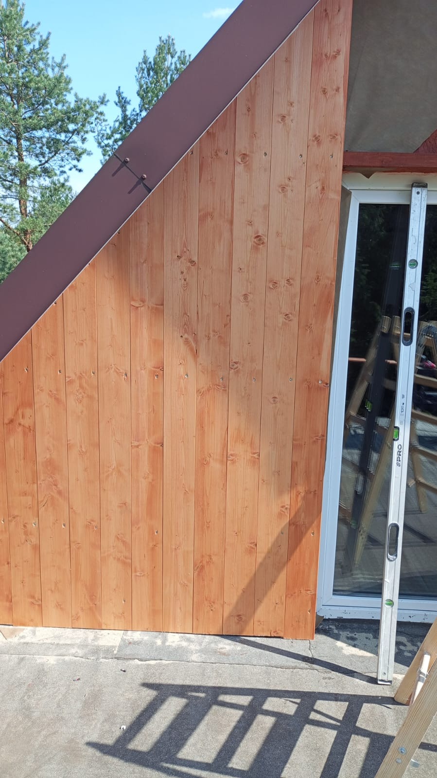 Drewniana elewacja domu w trakcie montażu, widoczne deski i poziomica przy oknie. Ciemny, skośny element dachu. Na dole cień drabiny.