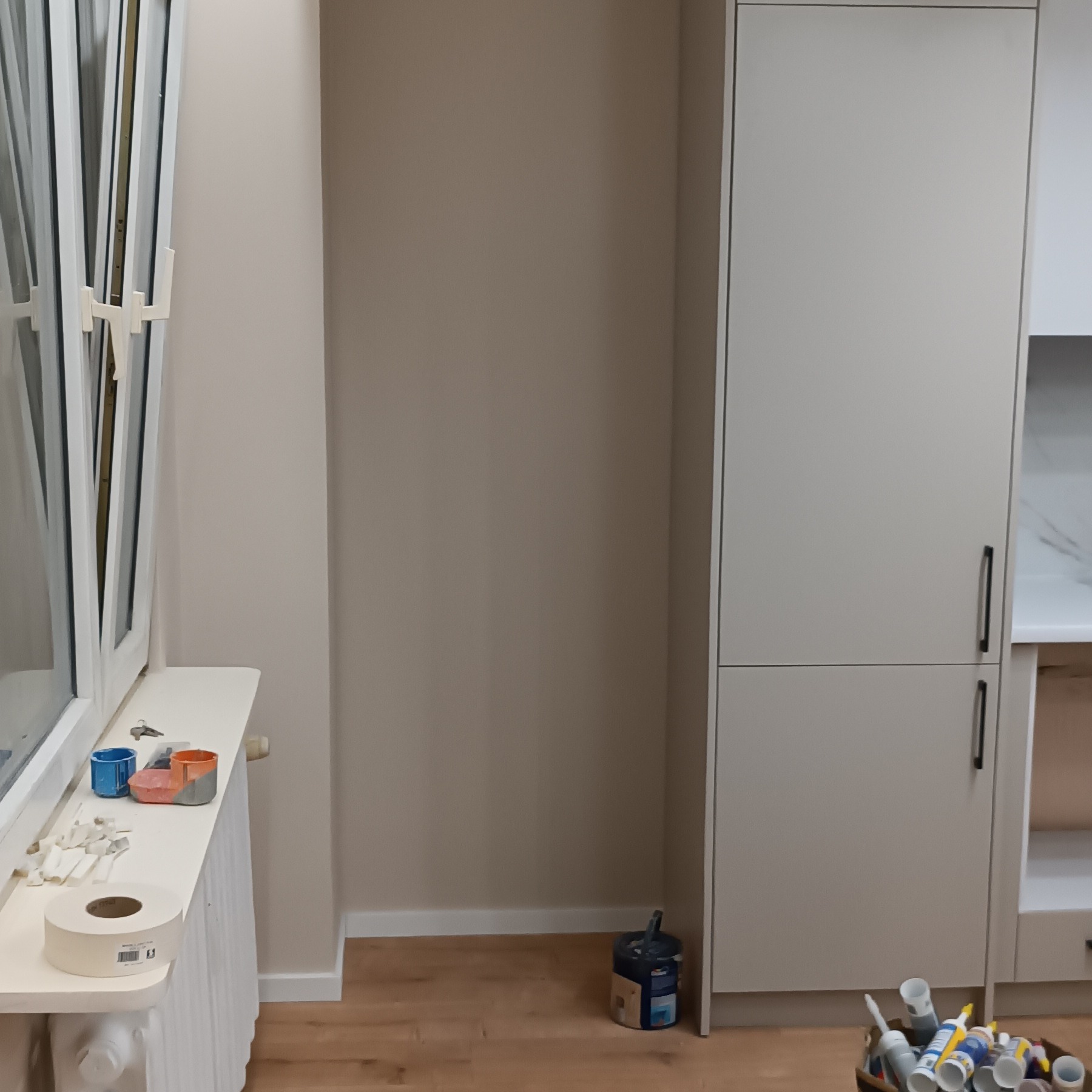 Wnętrze w trakcie prac wykończeniowych: ściany w neutralnym odcieniu beżu, zabudowa kuchenna, okno z akcesoriami malarskimi na parapecie i wiadro z narzędziami.