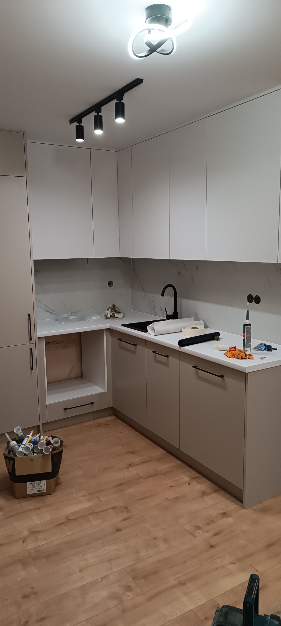 Jasna kuchnia z marmurowym blatem i czarną armaturą. Widoczne narzędzia i materiały montażowe, podkreślające etap wykończenia. Na suficie nowoczesne oświetlenie.