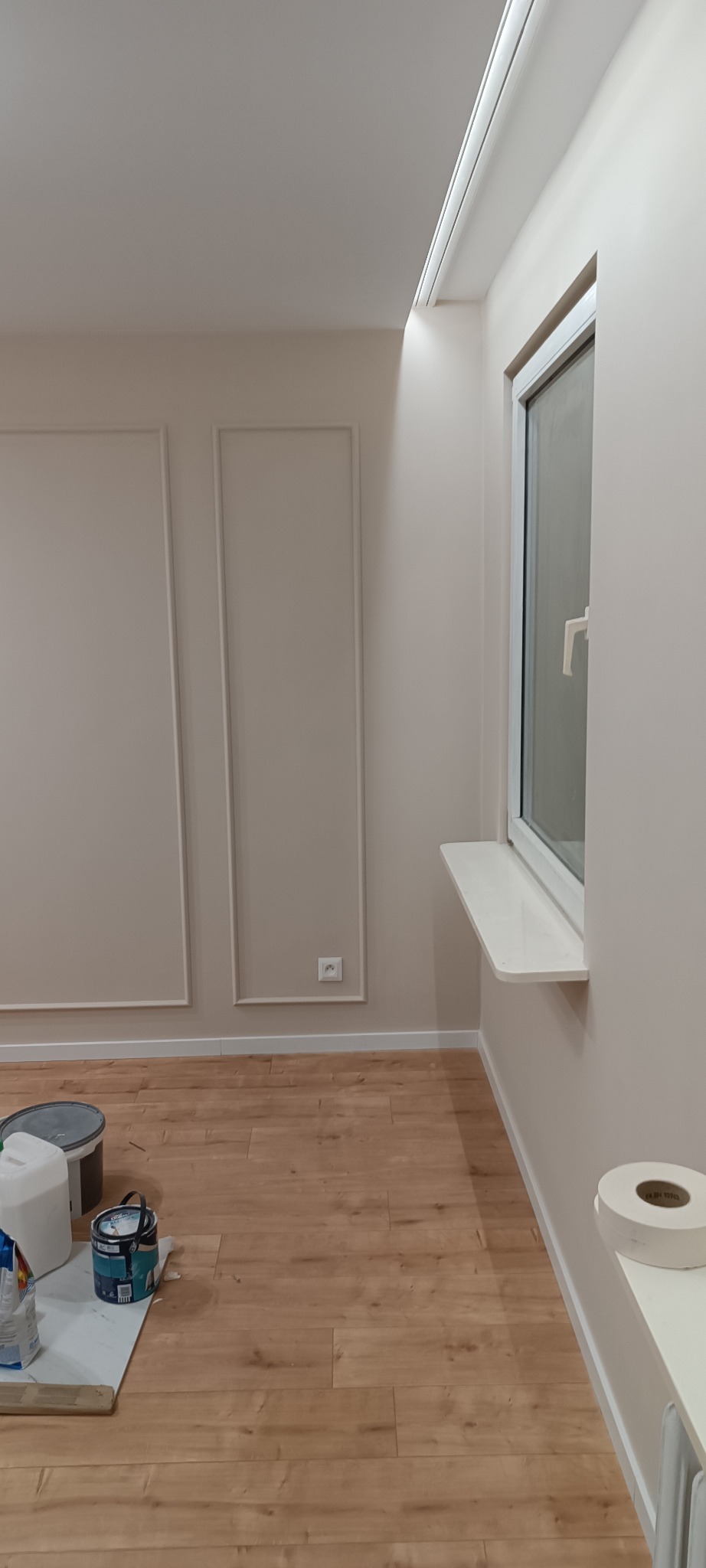 Jasne wnętrze po malowaniu ścian z dekoracyjnymi listwami i drewnopodobną podłogą. Widoczne puszki z farbą i akcesoria malarskie.