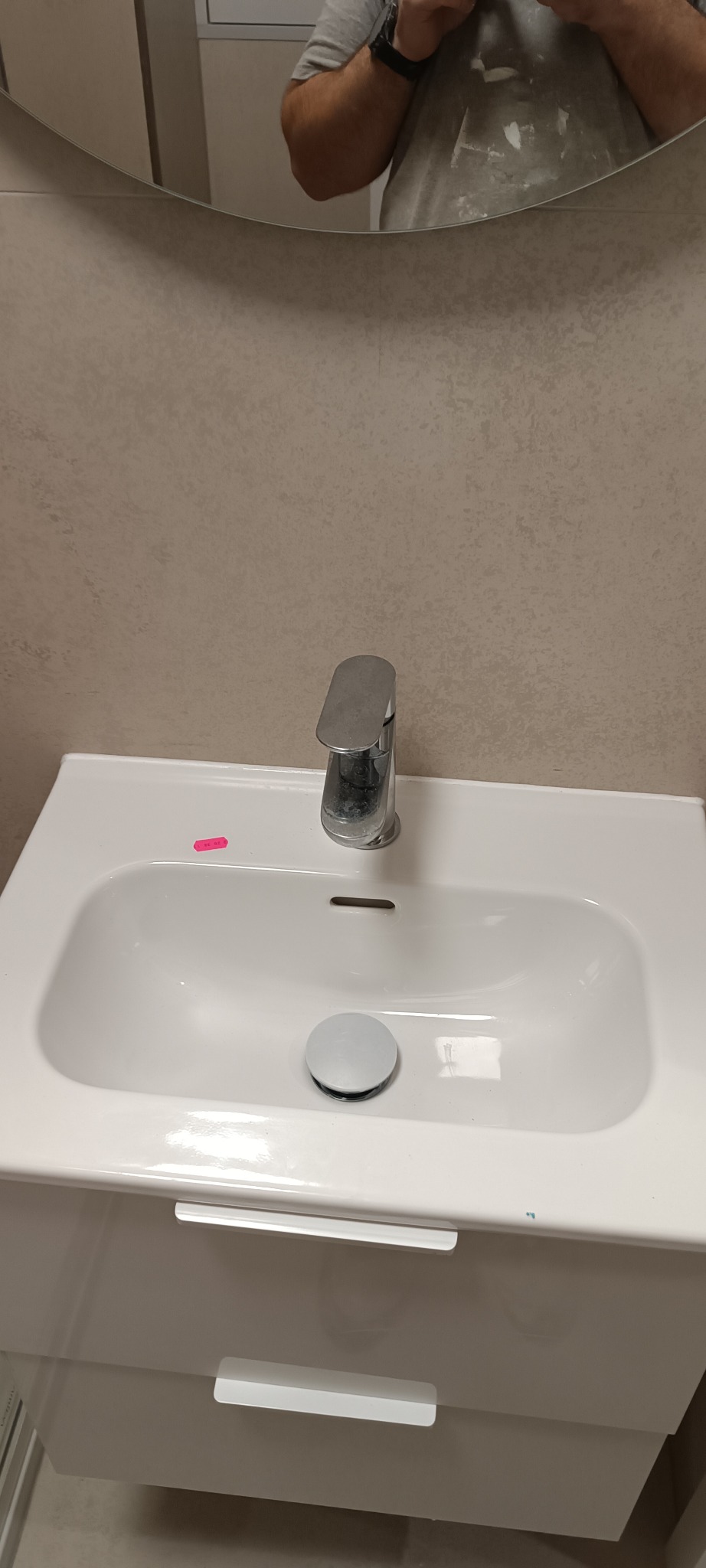 Biała umywalka z szafką, chromowana bateria. Na umywalce różowa karteczka. Widoczne odbicie osoby w lustrze nad umywalką. Neutralne tło.