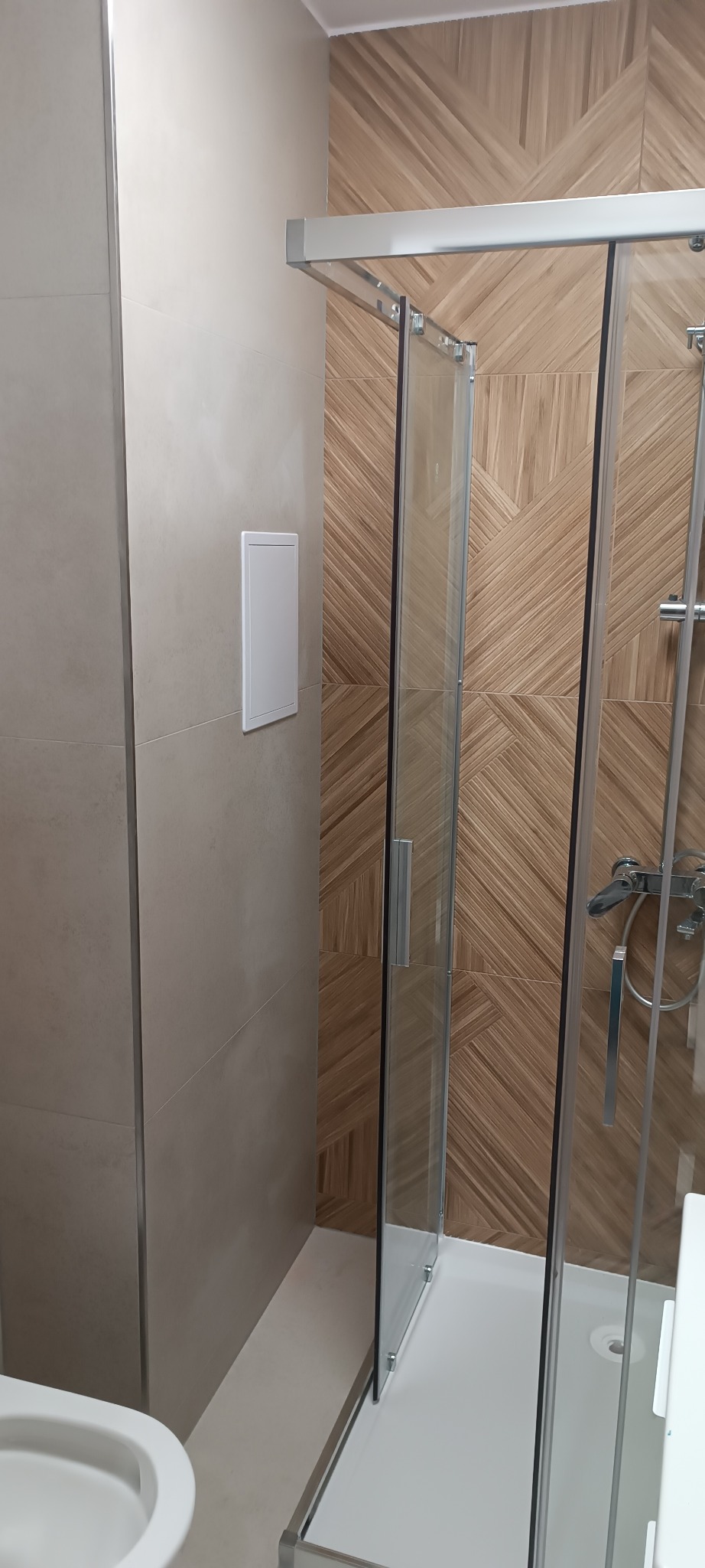 Nowoczesna kabina prysznicowa z drewnopodobnymi płytkami ułożonymi w geometryczny wzór. Widoczny biały panel rewizyjny na ścianie i fragment toalety.
