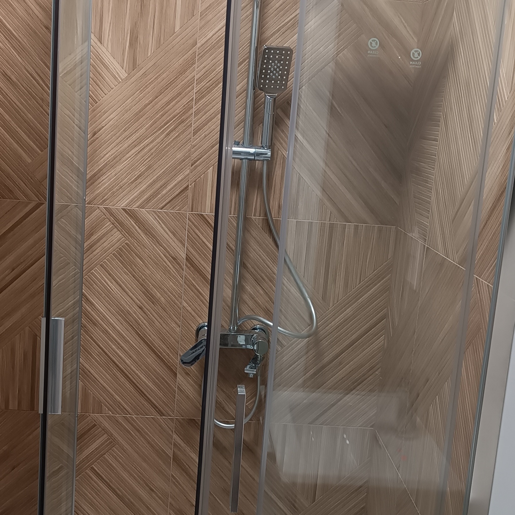 Kabina prysznicowa z deszczownicą na tle płytek imitujących drewno ułożonych w geometryczny wzór. Widoczne detale armatury i uchwyt drzwi.