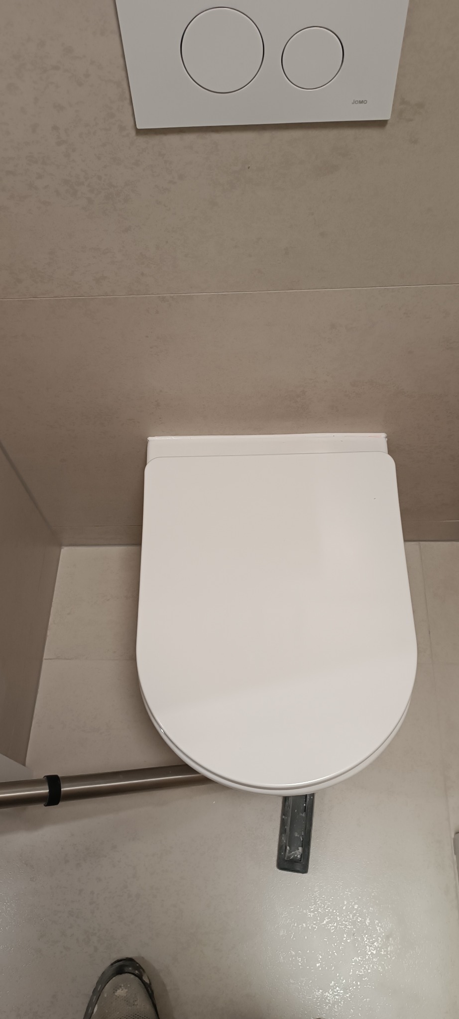 Nowoczesna toaleta wisząca JOMO z beżowymi płytkami w tle. Minimalistyczny design, widoczny fragment stopy w bucie roboczym. Ukończona instalacja w łazience.