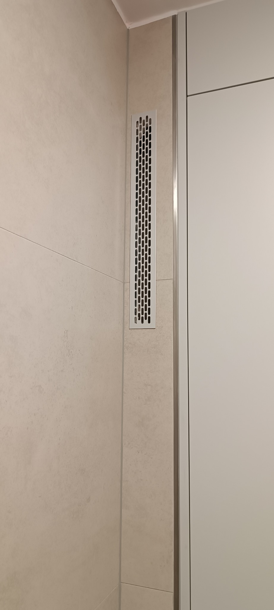 Wykończona łazienka: beżowe płytki ścienne z długą, wąską kratką wentylacyjną, sąsiadujące z białą zabudową meblową. Minimalistyczny design.