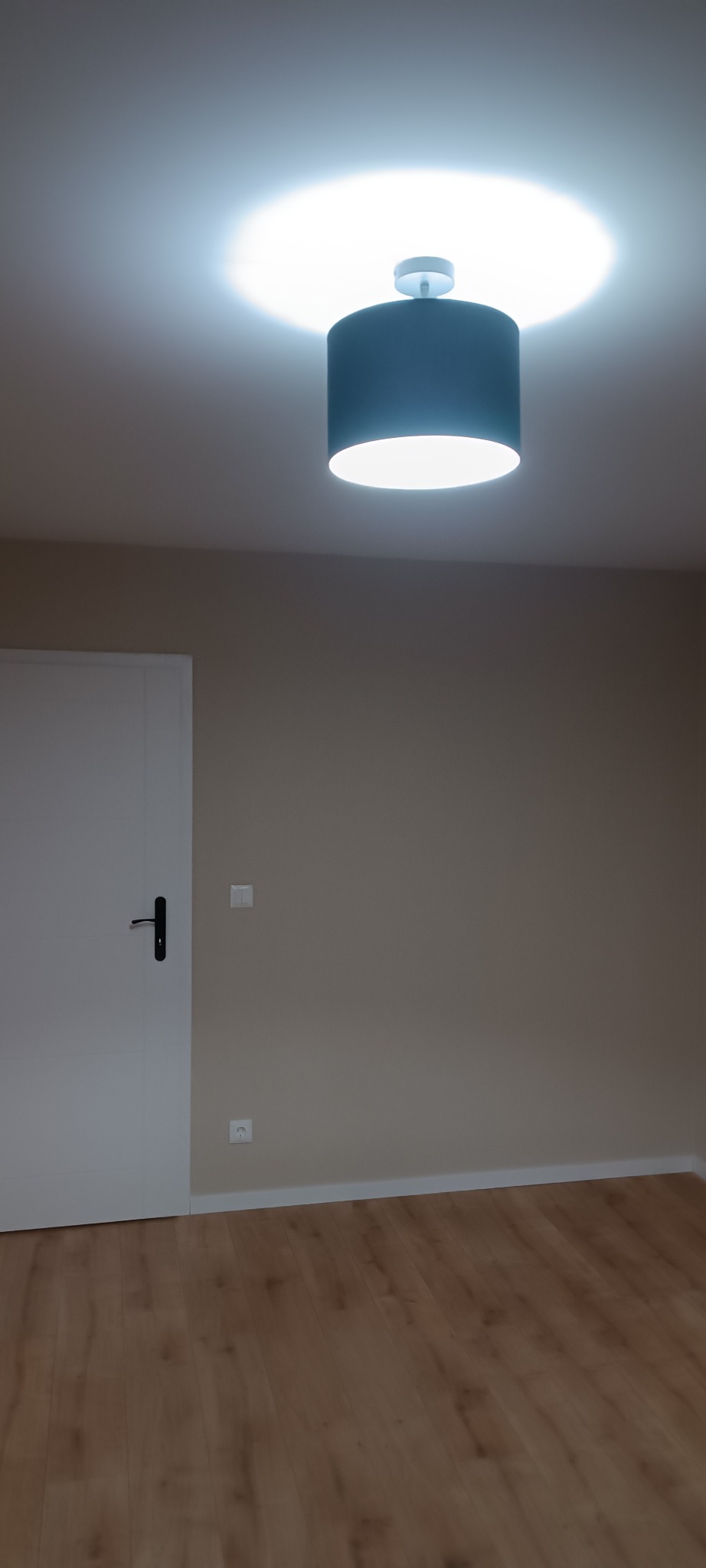 Pokój po remoncie: parkiet, ściany w kolorze beżowym, białe drzwi z czarną klamką i nowoczesna lampa sufitowa z abażurem w kolorze granatowym.