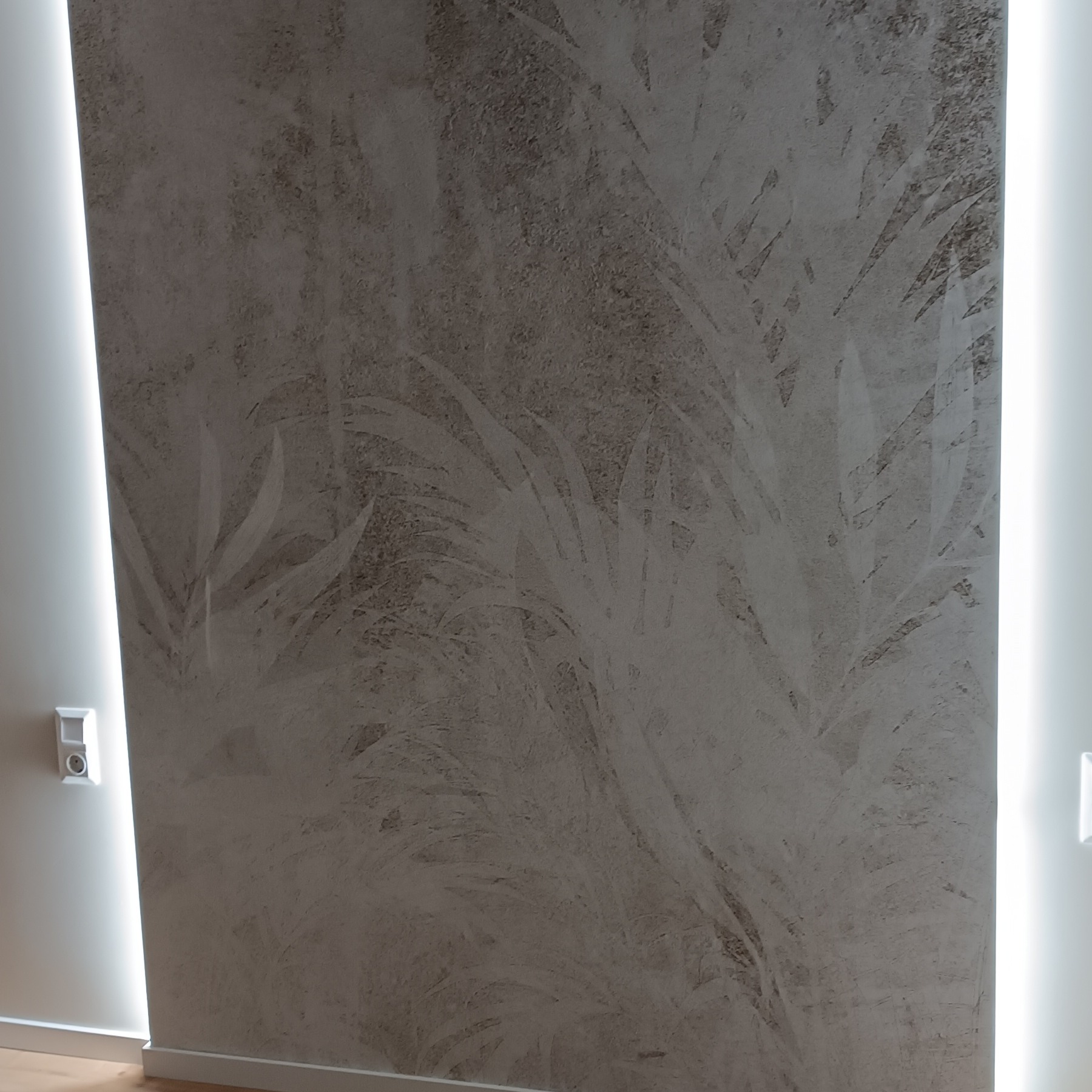Ściana z dekoracyjną, szarą fakturą przypominającą liście, oświetlona pionowymi pasami światła LED po bokach. Widoczny fragment podłogi i gniazdko elektryczne.