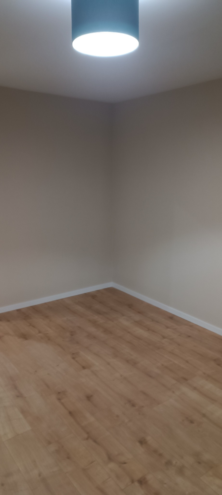 Jasne panele podłogowe w narożniku pokoju z beżowymi ścianami, oświetlone przez okrągłą lampę sufitową. Widoczne listwy przypodłogowe.