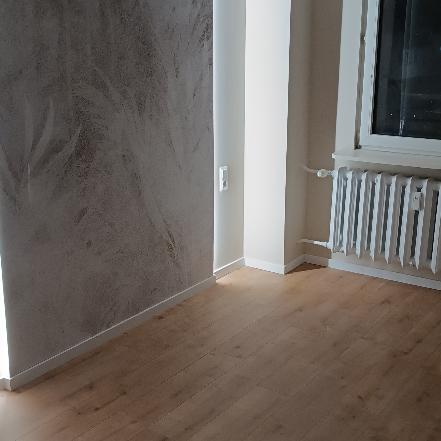 Wykończone wnętrze z dekoracyjną szarą ścianą, drewnianą podłogą i białym grzejnikiem pod oknem. Widoczny fragment ściany z gniazdkiem.