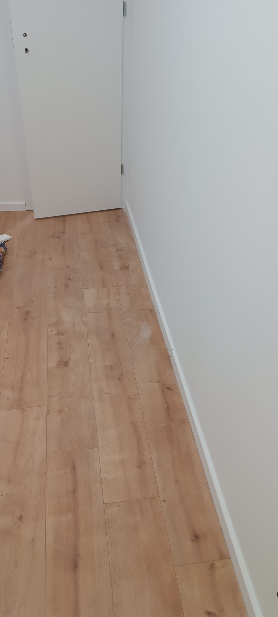 Jasne panele podłogowe ułożone wzdłuż białej ściany z listwą przypodłogową. W tle białe drzwi. Widoczny fragment podłogi i ściany po wykończeniu.