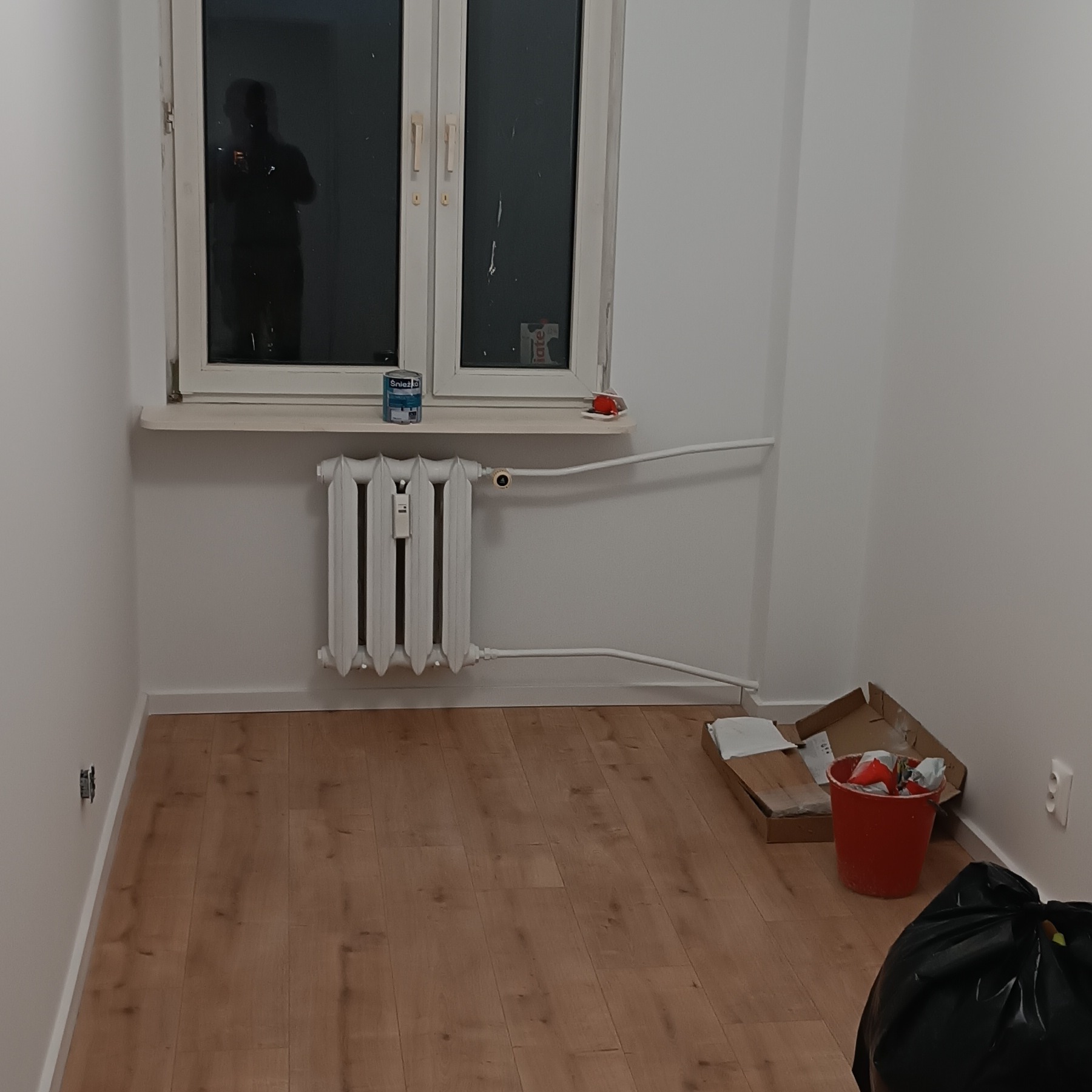 Pokój po malowaniu ścian w Augustowie. Widoczny grzejnik, okno z odbiciem osoby, podłoga z panelami oraz ślady po remoncie: puszka farby, karton i wiadro.