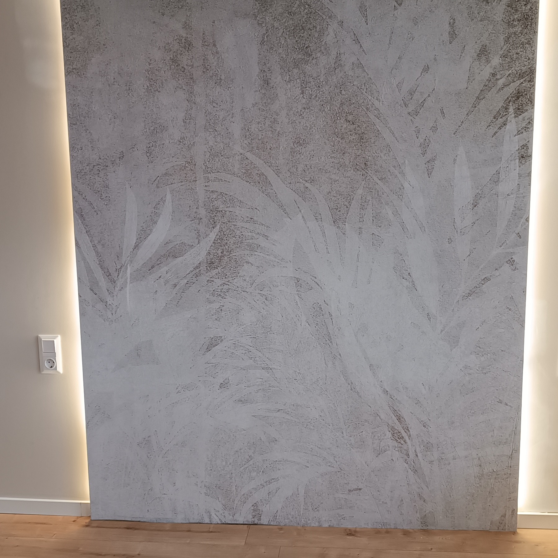 Dekoracyjna ściana z efektem betonu i motywem roślinnym, oświetlona pionowymi paskami LED, z widocznym gniazdkiem elektrycznym po lewej stronie.