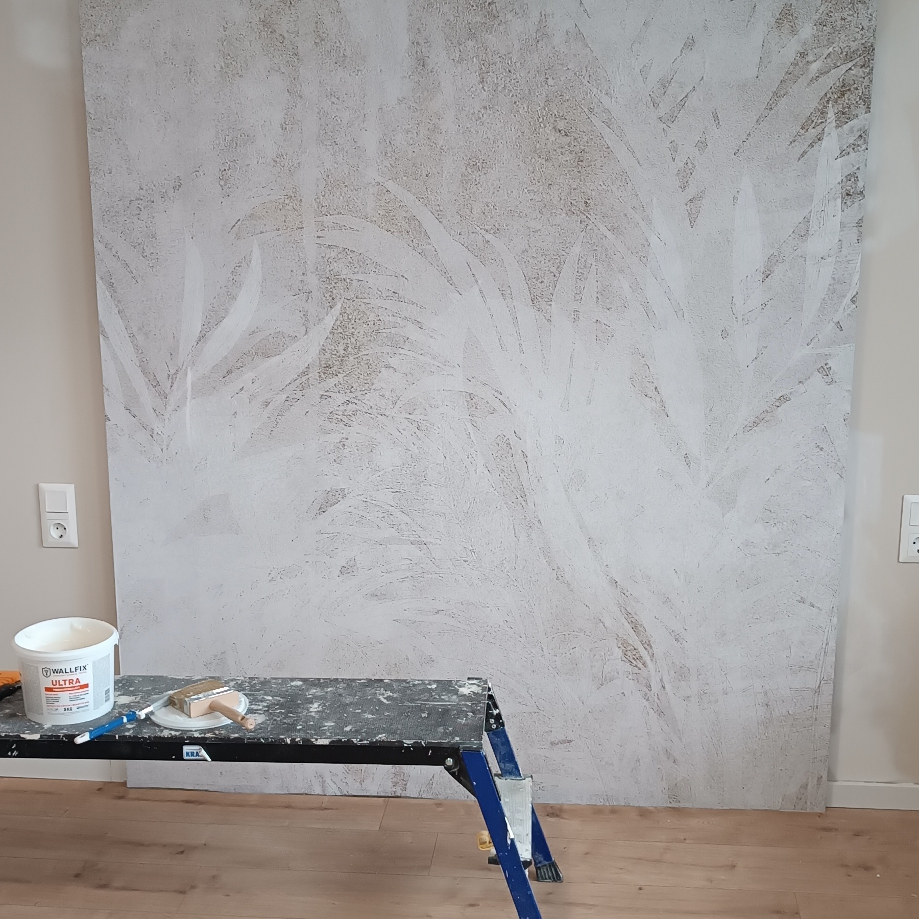 Ściana z dekoracyjną strukturą, częściowo pokryta białą farbą imitującą roślinny wzór. Obok drabina malarska z farbą i pędzlem.