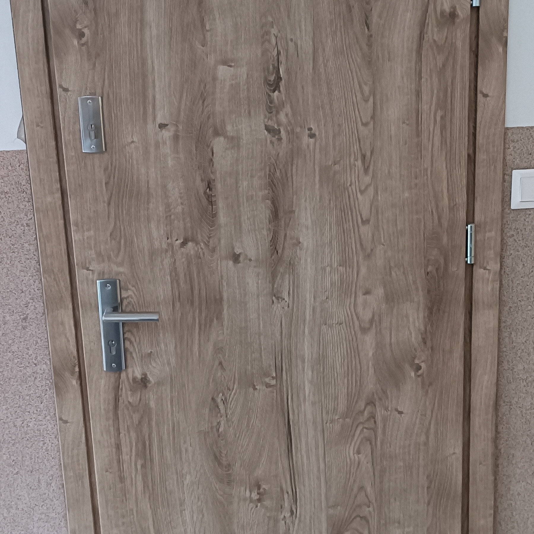 Zamknięte, drewnopodobne drzwi wewnętrzne z metalową klamką i zamkiem, widoczne na tle ściany pokrytej tapetą o drobnej fakturze. Oświetlenie dzienne.