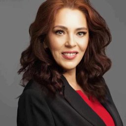 ALDAN NIERUCHOMOŚCI Alicja Danlowska - Portret uśmiechniętej kobiety z rudymi włosami w czarnej marynarce i czerwonej bluzce na szarym tle.