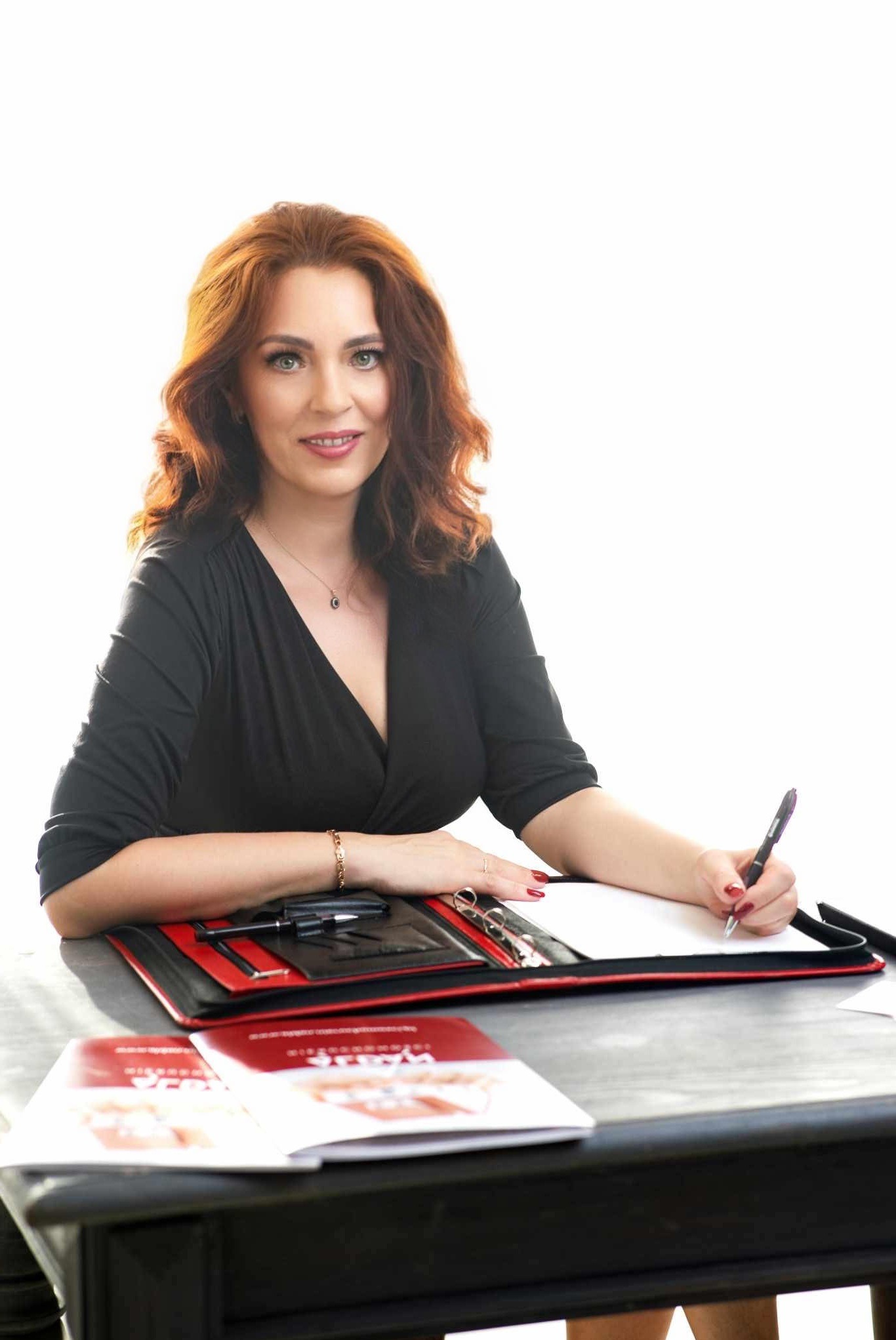 Kobieta w Zielonej Górze, uśmiechnięta, z długimi rudymi włosami, pisze w notatniku, obok leżą foldery reklamowe. Ujęcie portretowe, z bliska.