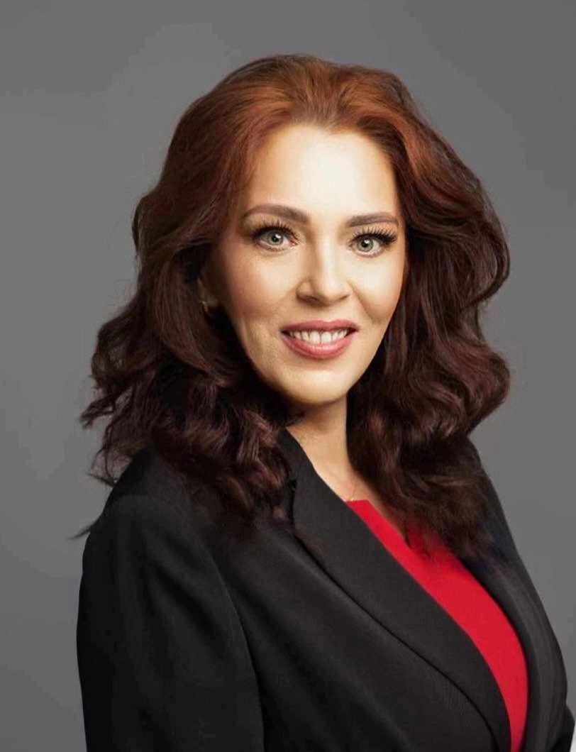 Portret uśmiechniętej kobiety z rudymi włosami w czarnej marynarce i czerwonej bluzce na szarym tle.