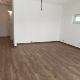 Puste, jasne pomieszczenie z panelami podłogowymi w odcieniach brązu, białe ściany z widocznymi gniazdkami i wystającym kablem, drzwi w tle.