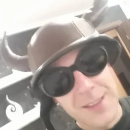 Uśmiechnięty mężczyzna w hełmie wikinga i okularach ochronnych pozuje na tle marmurowej ściany, z widocznymi fragmentami drzwi w tle.