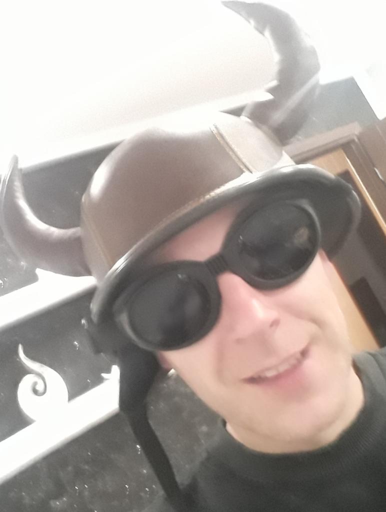 Uśmiechnięty mężczyzna w hełmie wikinga i okularach ochronnych pozuje na tle marmurowej ściany, z widocznymi fragmentami drzwi w tle.