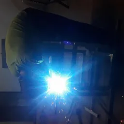 Pracownik w kombinezonie spawalniczym łączy metalowe elementy konstrukcji za pomocą spawarki, widoczne intensywne światło i iskry podczas spawania.