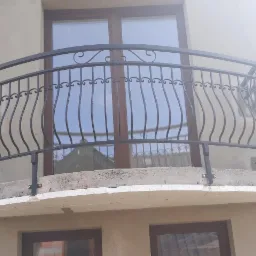 Metalowa balustrada balkonowa z dekoracyjnymi elementami, zamontowana na balkonie z widoczną warstwą ocieplenia, na tle brązowych ram okiennych.