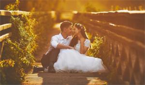 Para młoda w białej sukni i koszuli całuje się siedząc na drewnianym moście otoczonym zielenią w ciepłym, słonecznym świetle.