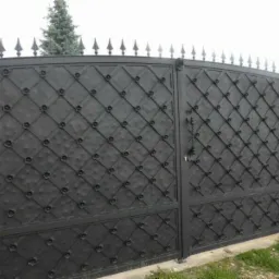 Czarna, dwuskrzydłowa brama wjazdowa z ozdobnym, romboidalnym wzorem i kwiatowymi detalami, zwieńczona ostrymi grotami, zamontowana między kamiennymi słupkami.