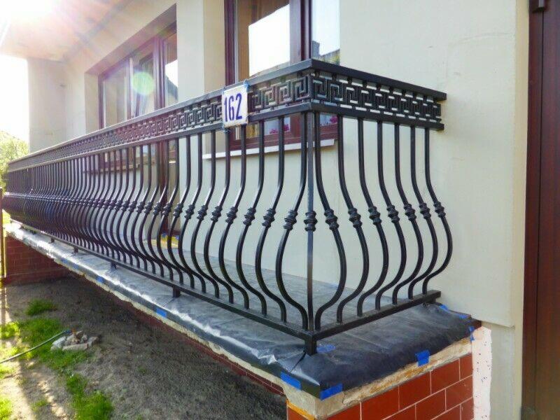 Czarna, metalowa balustrada balkonowa z motywem greckim na górze i numerem domu '162' przymocowanym do boku, widoczna folia ochronna na cokole.