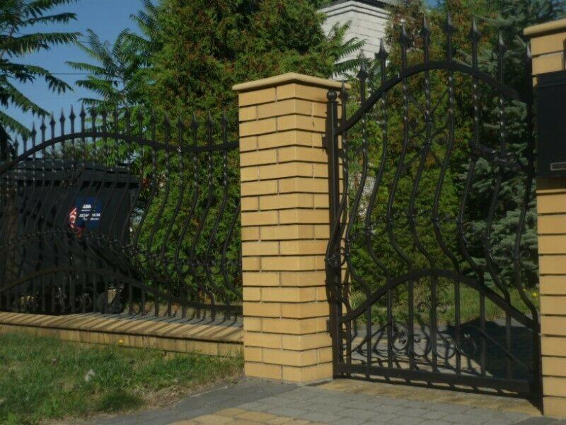 Kuta, czarna brama wjazdowa z dekoracyjnymi elementami, osadzona między dwoma słupkami z żółtej cegły, widok z zewnątrz na tle zieleni.