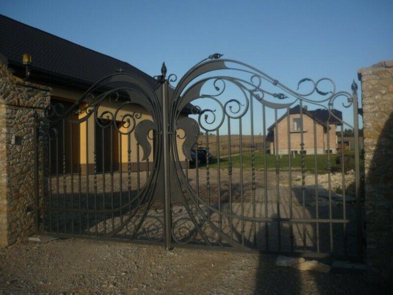 Ozdobna, metalowa brama wjazdowa z motywami roślinnymi, zamontowana między kamiennymi słupkami, widok na dom i podjazd.
