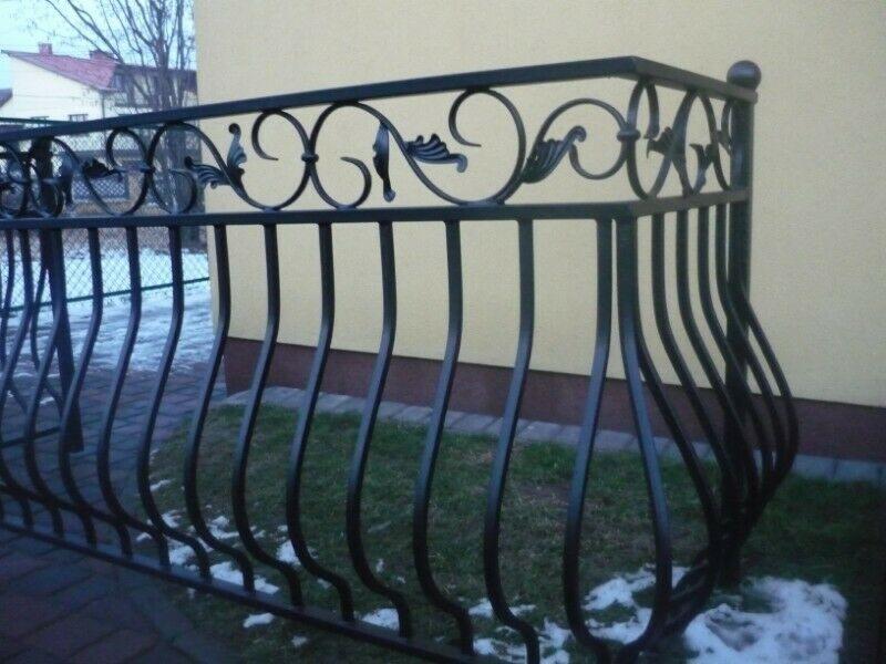 Kute, metalowe ogrodzenie z ozdobnymi elementami w kształcie liści i okręgów, widoczne na tle trawnika z resztkami śniegu i elewacji budynku.