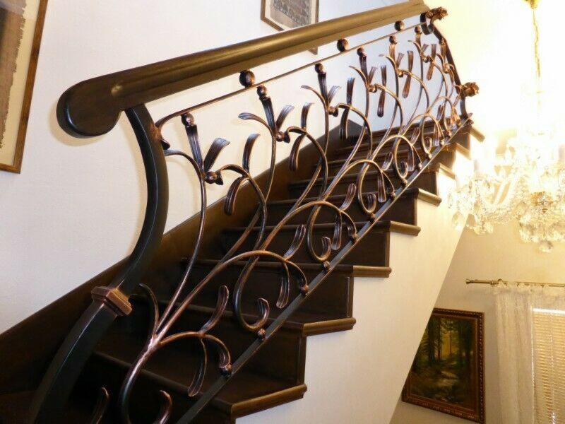 Kute, dekoracyjne balustrady w kolorze miedzi z motywem roślinnym, zamontowane przy drewnianych schodach w jasnym wnętrzu z kryształowym żyrandolem w tle.