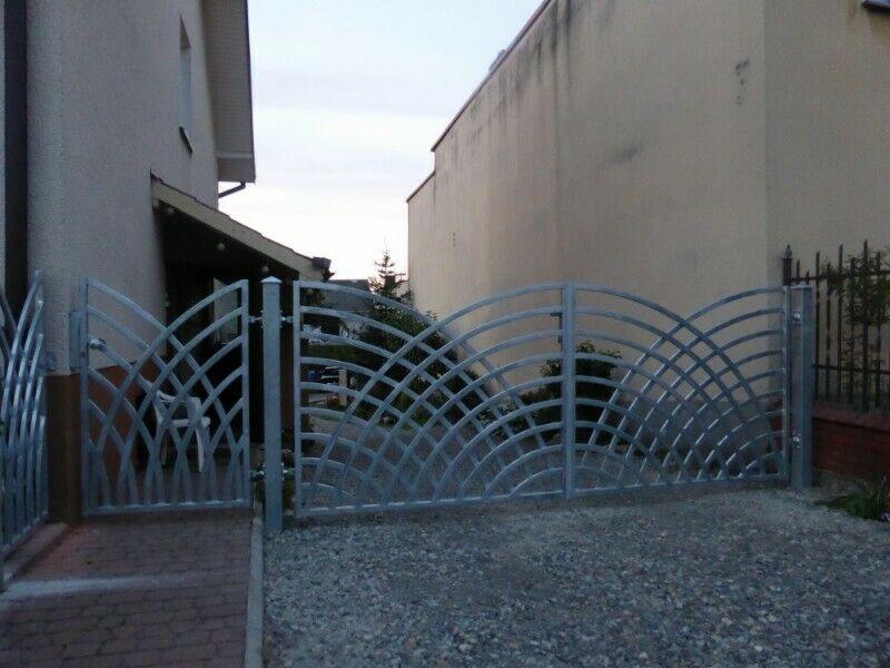 Metalowa brama dwuskrzydłowa z dekoracyjnym, ażurowym wypełnieniem w kształcie łuków, zamontowana na posesji z szarym żwirowym podłożem.