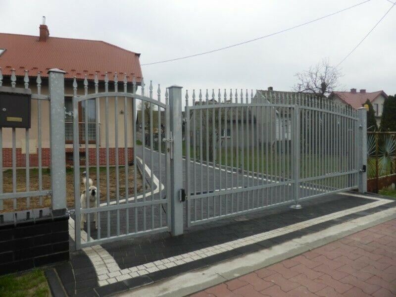 Szara, metalowa brama przesuwna z automatyką i furtka z widocznym psem rasy owczarek podhalański, na tle domu z czerwoną dachówką.