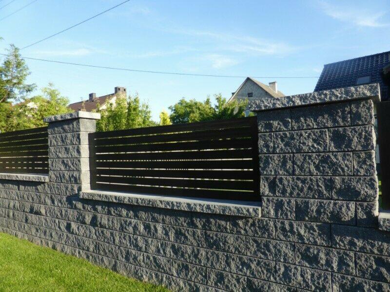 Nowoczesne ogrodzenie panelowe w kolorze czarnym, osadzone na murze z szarego kamienia łupanego, z widocznymi słupkami z tego samego materiału, na tle zielonego trawnika i zabudowań mieszkalnych.