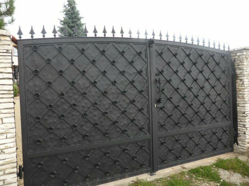 Czarna, dwuskrzydłowa brama wjazdowa z ozdobnym, romboidalnym wzorem i kwiatowymi detalami, zwieńczona ostrymi grotami, zamontowana między kamiennymi słupkami.