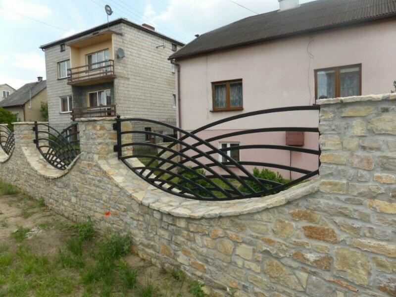 Kamienny murek z czarnym, metalowym, dekoracyjnym przęsłem ogrodzeniowym o falistym wzorze na tle dwóch budynków.