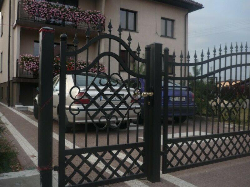 Czarna, metalowa brama wjazdowa z ozdobnymi elementami, widok na dom z balkonami obsadzonymi kwiatami i dwa samochody w tle.