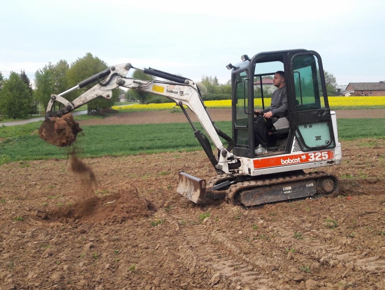 Operator koparki Bobcat 325 nabiera ziemię na polu uprawnym z rzepakiem w tle.