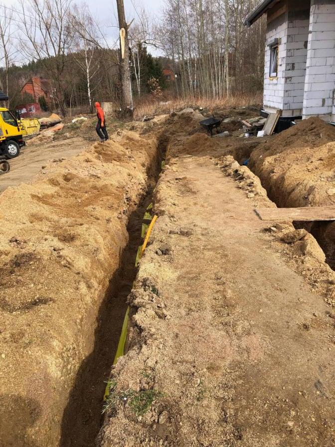 Wykop pod instalacje w ziemi przy budowie domu jednorodzinnego, widoczny żółty przewód w wykopie, na drugim planie pracownik w pomarańczowej kurtce i żółta koparka.