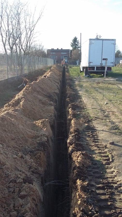 Wykopany rów w ziemi, widoczne ślady opon, w tle budynek mieszkalny i zaparkowana ciężarówka dostawcza.