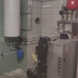 Kotłownia w trakcie montażu: biały zasobnik ciepłej wody, szary piec na pellet marki Metal-Fach Smart Pellet, czerwony zbiornik wyrównawczy ciśnienia, instalacja rurowa na ścianie z białych płytek,...