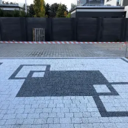 Ułożona kostka brukowa w nowoczesnym wzorze geometrycznym z szarych i białych kostek, z widoczną taśmą ostrzegawczą i paletą w tle, na tle ciemnego ogrodzenia i domu.